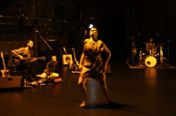 In der Mitte der Bühne ist eine Tänzerin. Hinter ihr sind ein Musiker und eine Schlagzeugerin. Eine andere Tänzerin sitzt auf dem Boden und schaut zu.  In der Mitte der Bühne ist eine Tänzerin. Hinter ihr sind ein Musiker und eine Schlagzeugerin. Eine andere Tänzerin sitzt auf dem Boden und schaut zu.