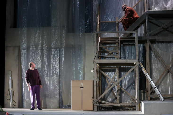 Ein mehrstöckiges Holzgerüst steht auf der Bühne. Ganz oben sitzt ein Schauspieler auf der Treppe. Er spricht mit einem anderen Schauspieler unten.  Ein mehrstöckiges Holzgerüst steht auf der Bühne. Ganz oben sitzt ein Schauspieler auf der Treppe. Er spricht mit einem anderen Schauspieler unten.