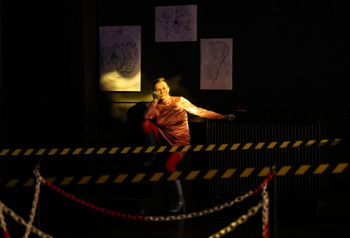 Vorn im Bild sind zwei Absperrbänder zu sehen. Hinten an der Wand sitzt eine Schauspielerin. Sie hat ihren Kopf in eine Hand gestützt. Vorn im Bild sind zwei Absperrbänder zu sehen. Hinten an der Wand sitzt eine Schauspielerin. Sie hat ihren Kopf in eine Hand gestützt.