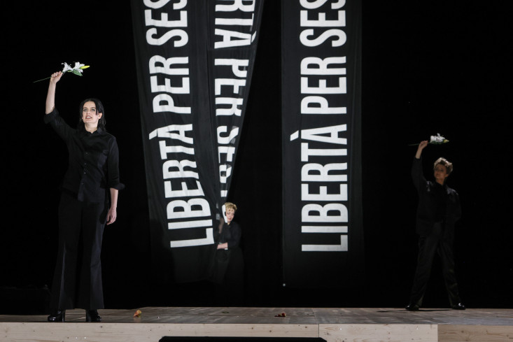 Zwei Sängerinnen stehen an den Bühnenseiten. Sie halten weiße Blumen hoch. In der Mitte der Bühne hängen drei Banner. Auf denen steht "Libertà per", mehr ist nicht zu lesen. 