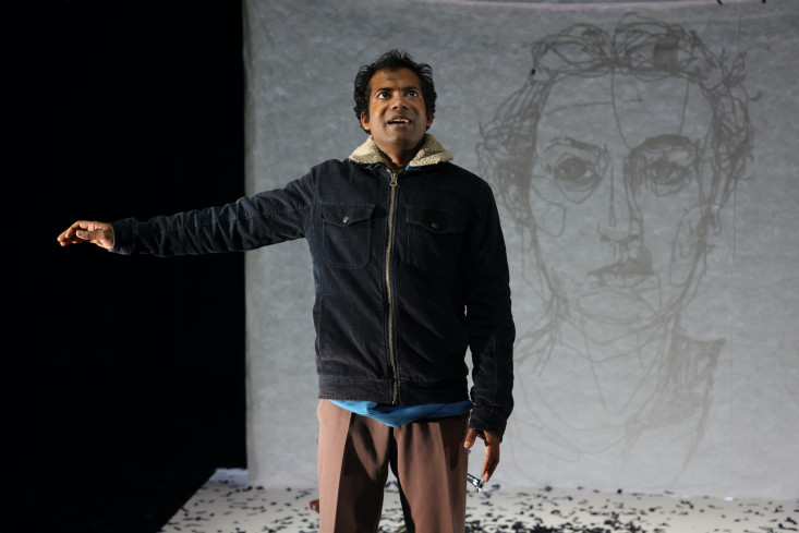 Der Schauspieler steht in Winterjacke in der Bühnenmitte und blickt nach oben. Sein rechter Arm ist ausgestreckt, als würde er einem Kind übers Haar streichen. Hinter ihm ist auf einer Leinwand das kunstvoll gezeichnete Porträt einer Frau zu sehen. Der Schauspieler steht in Winterjacke in der Bühnenmitte und blickt nach oben. Sein rechter Arm ist ausgestreckt, als würde er einem Kind übers Haar streichen. Hinter ihm ist auf einer Leinwand das kunstvoll gezeichnete Porträt einer Frau zu sehen.