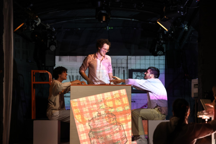 Eine Person steht hinter einer hüfthohen Kulisse und lächelt. Zwei andere Menschen sitzen rechts und links davon. Eine Person steht hinter einer hüfthohen Kulisse und lächelt. Zwei andere Menschen sitzen rechts und links davon.