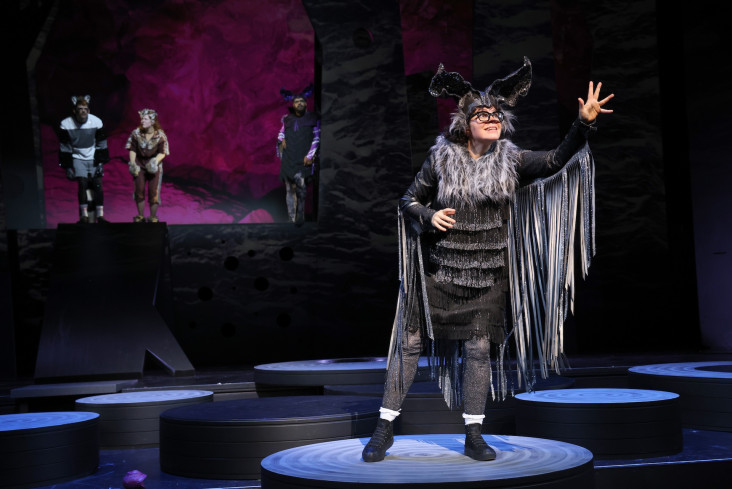 Eine Schauspielerin in einem Fledermauskostüm steht vorn auf der Bühne. Sie wird von drei anderen Personen, die weiter hinten stehen, beobachtet. Eine Schauspielerin in einem Fledermauskostüm steht vorn auf der Bühne. Sie wird von drei anderen Personen, die weiter hinten stehen, beobachtet.