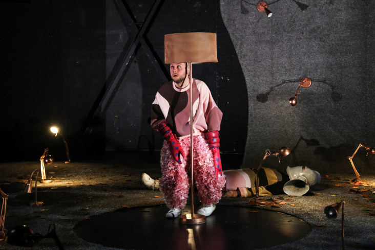 Ein Schauspieler duckt sich hinter einer Stehlampe. Er trägt eine rosane, bauschige Hose und Krebsscheren an den Händen. Ein Schauspieler duckt sich hinter einer Stehlampe. Er trägt eine rosane, bauschige Hose und Krebsscheren an den Händen.