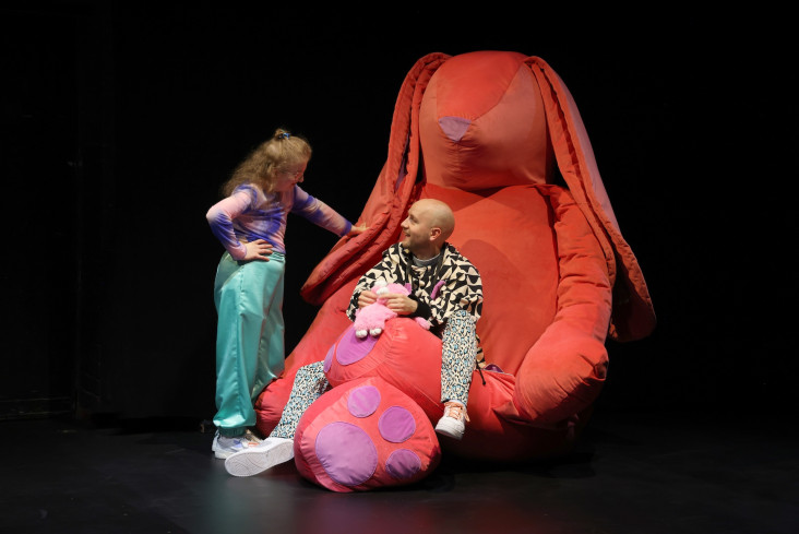 Auf dem Schoß eines riesigen, roten Stoffhasens sitzt ein Schauspieler. Neben ihm steht eine Schauspielerin. Er lächelt sie an. Auf dem Schoß eines riesigen, roten Stoffhasens sitzt ein Schauspieler. Neben ihm steht eine Schauspielerin. Er lächelt sie an.