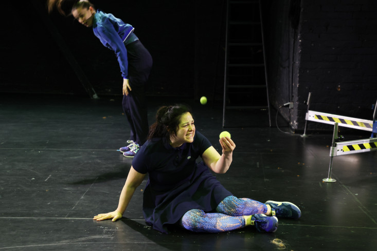 Eine Schauspielerin sitzt auf dem Boden. In der Hand hält sie einen Tennisball.