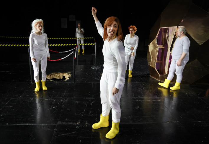 Fünf Personen sind auf der Bühne verteilt. Alle tragen weiße langärmelige Unterhemden und weiße, lange Unterhosen. Dazu gelbe Gummistiefel.