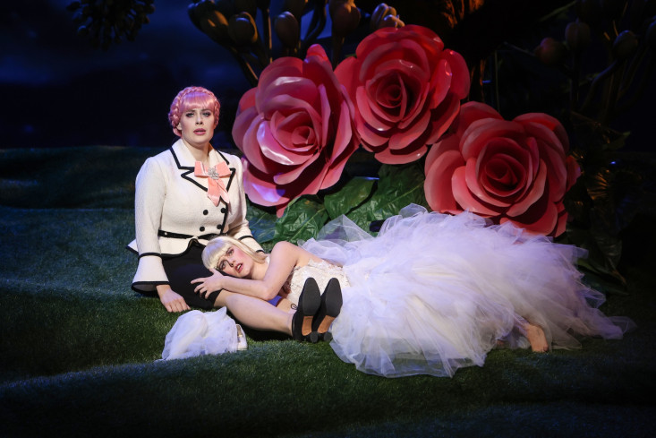Eine Sängerin in einem Brautkleid hat ihren Kopf in den Schoß einer anderen Sängerin gelegt. Hinter ihnen sind riesige, rote Rosen. Eine Sängerin in einem Brautkleid hat ihren Kopf in den Schoß einer anderen Sängerin gelegt. Hinter ihnen sind riesige, rote Rosen.