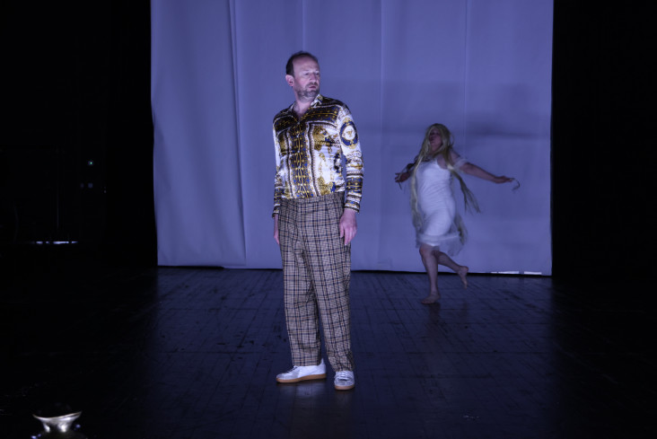 Ein Schauspieler steht vorn auf der Bühne. Hinter ihm tanzt eine Schauspielerin in einem weißen Kleid. Ein Schauspieler steht vorn auf der Bühne. Hinter ihm tanzt eine Schauspielerin in einem weißen Kleid.