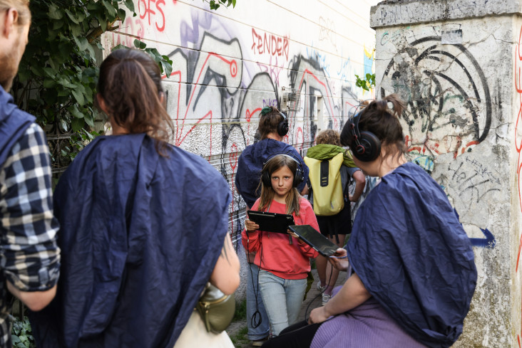 Eine Gruppe von Menschen mit Kopfhörern auf den Ohren und Tablets in den Händen. Sie stehen vor einer Wand irgendwo in der Stadt. Die Wand ist voller Graffiti. 