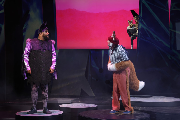 Auf der Bühne stehen zwei Schauspieler. Der eine trägt Fledermausohren, der andere Fuchsohren und einen Fuchsschwanz. Sie schauen sich an. Auf der Bühne stehen zwei Schauspieler. Der eine trägt Fledermausohren, der andere Fuchsohren und einen Fuchsschwanz. Sie schauen sich an.