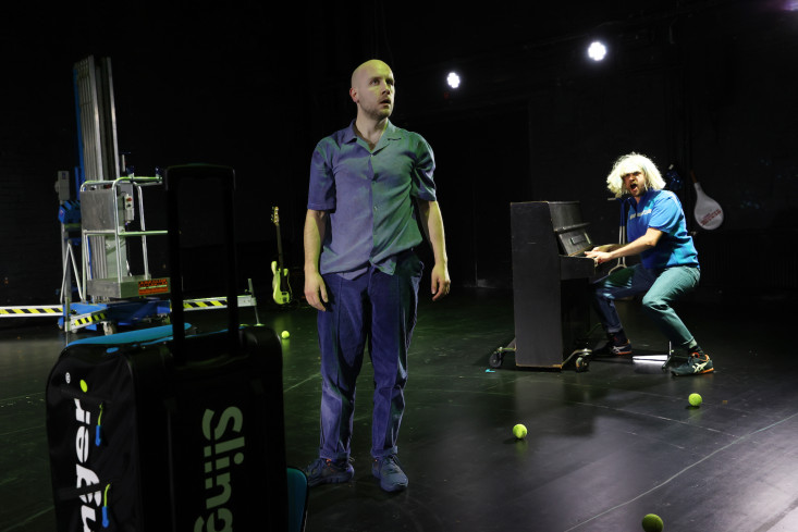 Ein Schauspieler steht dem Publikum zugewandt auf der Bühne. Hinten sitzt ein anderer Schauspieler mit einer blonden Langhaar-Perücke am Klavier. Ein Schauspieler steht dem Publikum zugewandt auf der Bühne. Hinten sitzt ein anderer Schauspieler mit einer blonden Langhaar-Perücke am Klavier.