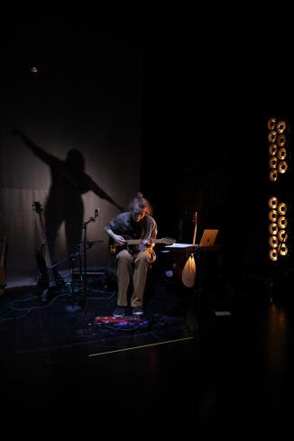 Ein Musiker sitzt auf einem Stuhl und spielt ein Saiteninstrument. Neben ihm stehen viele andere Instrumente. Auf der Leinwand hinter ihm ist sein eigener Schatten zu sehen. Ein Musiker sitzt auf einem Stuhl und spielt ein Saiteninstrument. Neben ihm stehen viele andere Instrumente. Auf der Leinwand hinter ihm ist sein eigener Schatten zu sehen.