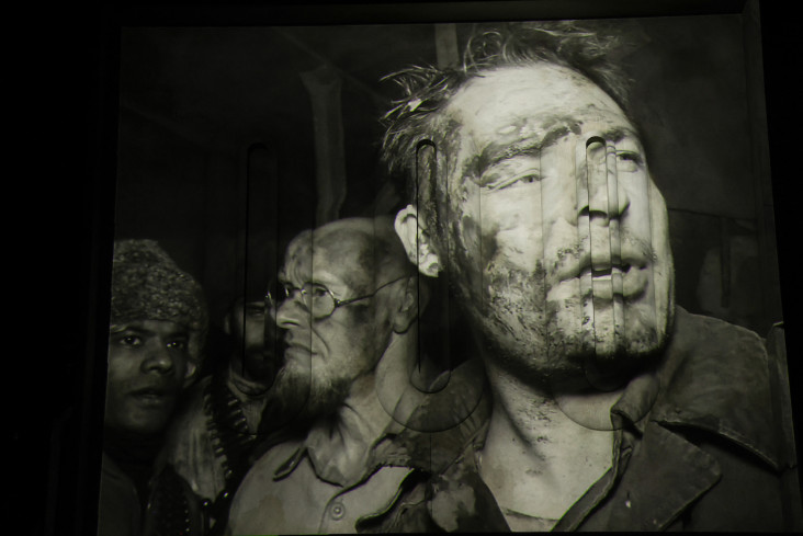 Eine Videoprojektion in schwarz und weiß zeigt drei Gesichter in Großaufnahme. Alle sind mit Schmutz oder Blut verschmiert. Eine Videoprojektion in schwarz und weiß zeigt drei Gesichter in Großaufnahme. Alle sind mit Schmutz oder Blut verschmiert.