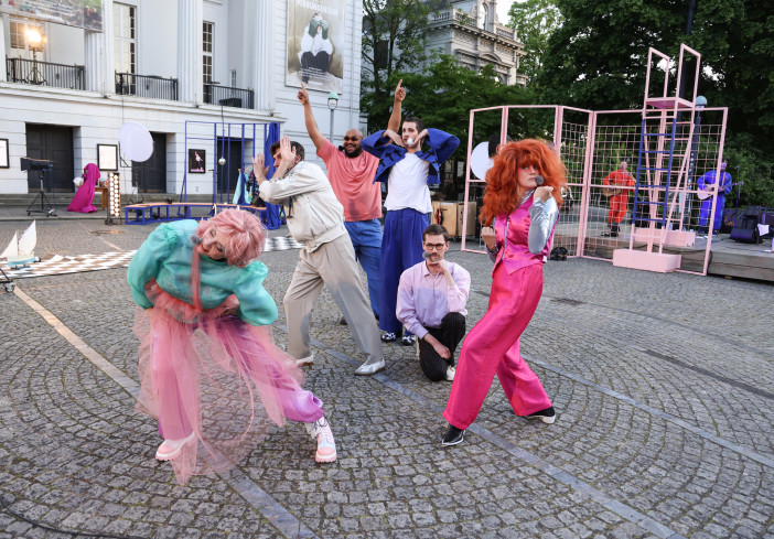 Sechs Personen stehen auf dem Goetheplatz. Fast alle sind sehr auffällig und farbenfroh gekleidet. Sie tanzen und singen. Im Hintergrund spielt eine Band. Sechs Personen stehen auf dem Goetheplatz. Fast alle sind sehr auffällig und farbenfroh gekleidet. Sie tanzen und singen. Im Hintergrund spielt eine Band.