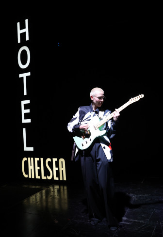 Eine Gitarristin steht im vollen Scheinwerferlicht. Neben ihr leuchtet ein Schild. Auf dem steht Hotel Chelsea. Eine Gitarristin steht im vollen Scheinwerferlicht. Neben ihr leuchtet ein Schild. Auf dem steht Hotel Chelsea.