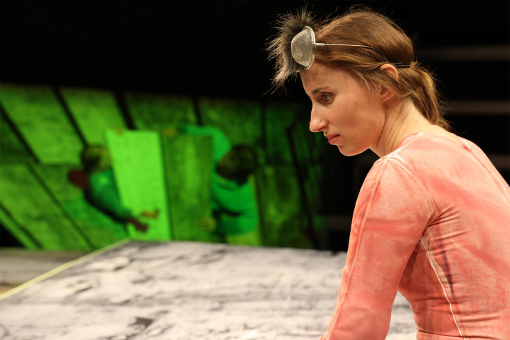 Eine Schauspielerin sitzt am Bühnenrand. Im Hintergrund sind zwei andere Personen hinter einer Klappe im Bühnenbild halb versteckt. Eine Schauspielerin sitzt am Bühnenrand. Im Hintergrund sind zwei andere Personen hinter einer Klappe im Bühnenbild halb versteckt.