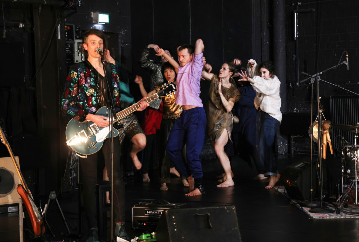 Ein Musiker singt und spielt E-Gitarre. Hinter ihm tanzen fünf Personen eng beieinander. Ein Musiker singt und spielt E-Gitarre. Hinter ihm tanzen fünf Personen eng beieinander.