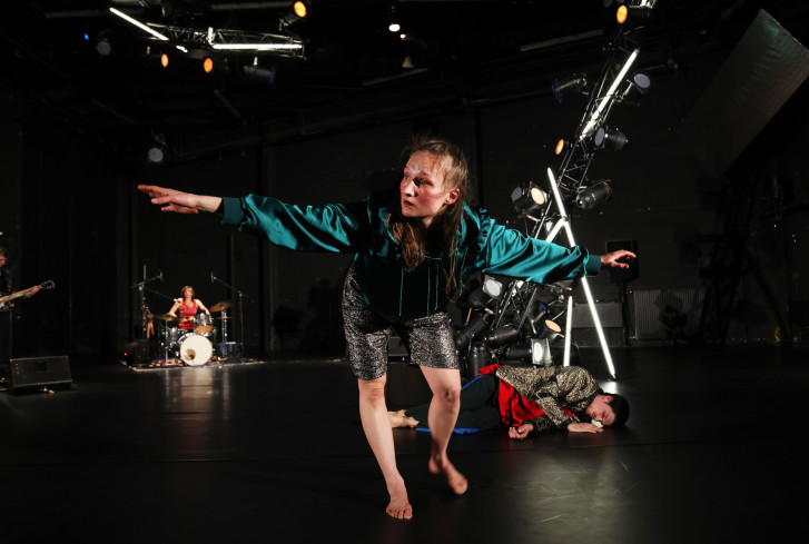 Eine Tänzerin steht vorgebeugt auf einem Bein. Sie hat die Arme ausgebreitet. Hinter ihr liegt ein Tänzer auf dem Boden. Eine Musikerin sitzt am Schlagzeug. Eine Tänzerin steht vorgebeugt auf einem Bein. Sie hat die Arme ausgebreitet. Hinter ihr liegt ein Tänzer auf dem Boden. Eine Musikerin sitzt am Schlagzeug.