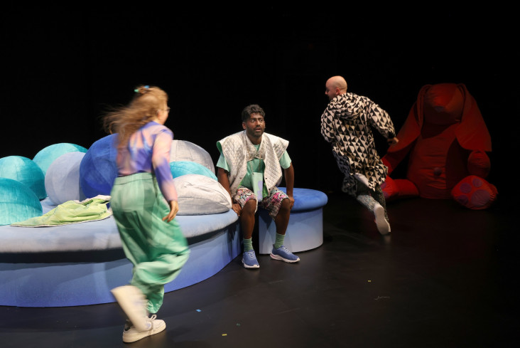 Ein Schauspieler sitzt auf einem mit Kissen bedeckten Podest mitten auf der Bühne. Zwei andere Personen laufen um ihn herum. Ein Schauspieler sitzt auf einem mit Kissen bedeckten Podest mitten auf der Bühne. Zwei andere Personen laufen um ihn herum.