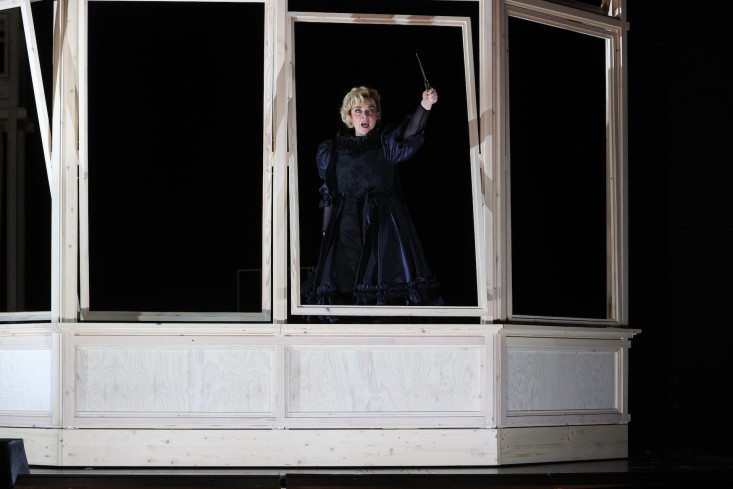 In einem Holzbau mit sehr großen Fenstern steht eine Sängerin. Die Bühne ist dunkel, sie ist schwarz gekleidet. Nur das Holz ist hell. 