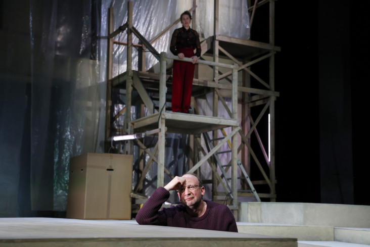 Vorn auf der Bühne sitzt ein Schauspieler. Er hat den Kopf in eine Hand gestützt. Hinten steht eine Schauspielerin auf einem Holzgerüst. Sie schaut ihn an.  Vorn auf der Bühne sitzt ein Schauspieler. Er hat den Kopf in eine Hand gestützt. Hinten steht eine Schauspielerin auf einem Holzgerüst. Sie schaut ihn an.