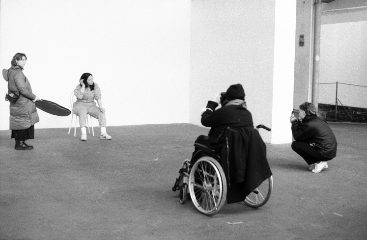 Eine Frau sitzt auf einem Hocker. Neben ihr steht eine andere Frau. Ihnen gegenüber sind zwei Personen, beide fotografieren. Eine sitzt in einem Rollstuhl.