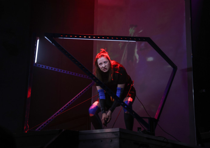 Eine Sängerin hockt auf einem Podest. Das Podest ist von zwei Eisengeländern umgeben. Im Hintergrund ist eine rötlich leuchtende Wand. 