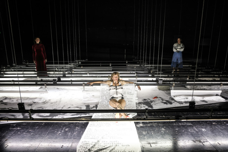 Auf der ganzen Bühne sind auf Kniehöhe lange Stangen. An ihnen hängen Schriftbahnen, die jetzt zerknüllt und nicht mehr lesbar sind. In der Mitte der Bühne sitzt eine Schauspielerin auf der einzigen ausgebreiteten Schriftbahn. Zwei Schauspielerinnen stehen im Hingergrund. Auf der ganzen Bühne sind auf Kniehöhe lange Stangen. An ihnen hängen Schriftbahnen, die jetzt zerknüllt und nicht mehr lesbar sind. In der Mitte der Bühne sitzt eine Schauspielerin auf der einzigen ausgebreiteten Schriftbahn. Zwei Schauspielerinnen stehen im Hingergrund.