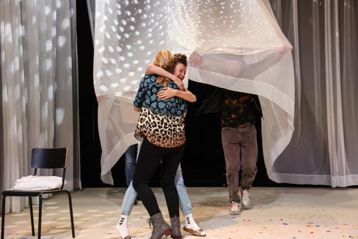 Zwei Schauspielerinnen umarmen sich. Hinter ihnen hängt ein Vorhang. Ein Mann hebt ihn an. Zwei Schauspielerinnen umarmen sich. Hinter ihnen hängt ein Vorhang. Ein Mann hebt ihn an.