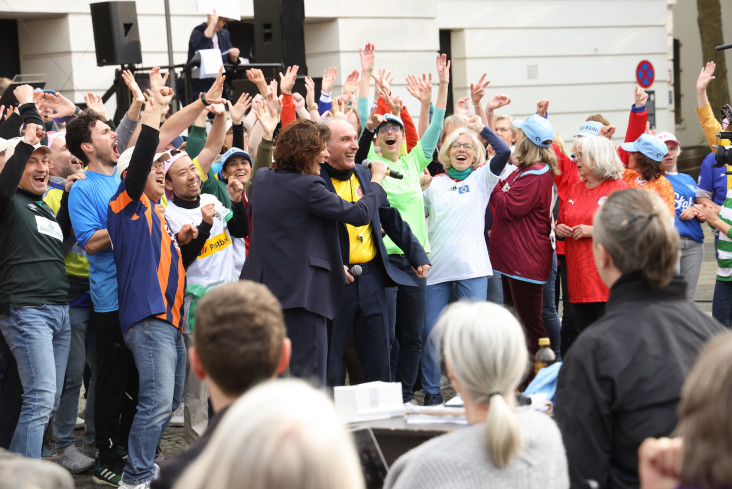 Sehr viele Menschen in Fußballtrikots stehen eng beieinander. In ihrer Mitte stehen zwei Personen. Die eine hält der anderen ein Mikrofon vor den Mund. Alle sind draußen vor dem Theater. Sehr viele Menschen in Fußballtrikots stehen eng beieinander. In ihrer Mitte stehen zwei Personen. Die eine hält der anderen ein Mikrofon vor den Mund. Alle sind draußen vor dem Theater.