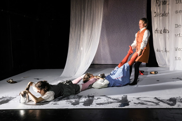 Eine Schauspielerin liegt auf dem Bauch. Ihre Hand steckt in einem Eimer mit Farbe. Ihre Beine werden von einer zweiten Schauspielerin festgehalten. Deren Beine wiederum von einer dritten gehalten werden. Eine Schauspielerin liegt auf dem Bauch. Ihre Hand steckt in einem Eimer mit Farbe. Ihre Beine werden von einer zweiten Schauspielerin festgehalten. Deren Beine wiederum von einer dritten gehalten werden.