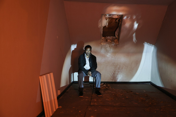 Ein Mann sitzt auf einem Stuhl in der hinteren Ecke des Raums. Auf der Wand hinter ihm läuft ein großes Video. Er selbst ist auf dem Video zu sehen. Ein Mann sitzt auf einem Stuhl in der hinteren Ecke des Raums. Auf der Wand hinter ihm läuft ein großes Video. Er selbst ist auf dem Video zu sehen.