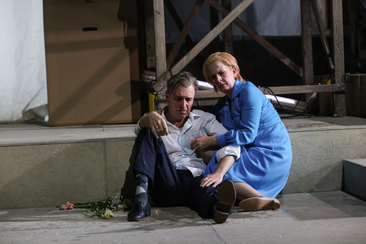 Eine Schauspielerin und ein Schauspieler sitzen nebeneinander auf dem Boden. Sie hat einen Arm um ihn gelegt. Er schaut auf seine Schuhe. 