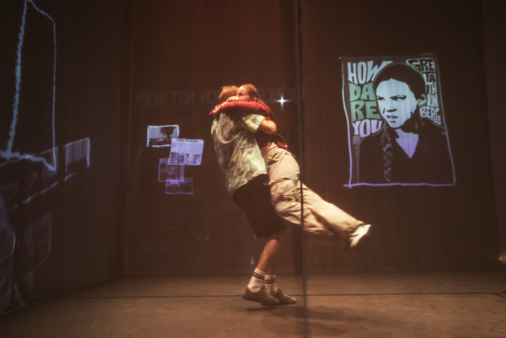 Ein Schauspieler schwenkt eine Schauspielerin durch die Luft. Im Hintergrund sind Bilder an die Wand projiziert. Vor der Szene hängt eine durchsichtige Gaze. Ein Schauspieler schwenkt eine Schauspielerin durch die Luft. Im Hintergrund sind Bilder an die Wand projiziert. Vor der Szene hängt eine durchsichtige Gaze.