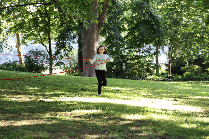 Eine junge Akteurin läuft über eine Wiese in einem Park. Sie hält sich an einem roten Seil fest.