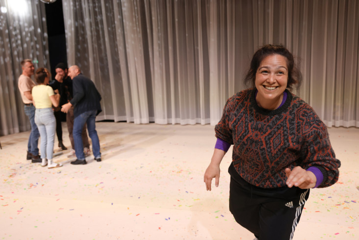Eine Schauspielerin steht vorn an der Bühnenkante. Sie ist dem Publikum zugewandt. Hinten in der Ecke stehen andere in einem Kreis. Eine Schauspielerin steht vorn an der Bühnenkante. Sie ist dem Publikum zugewandt. Hinten in der Ecke stehen andere in einem Kreis.