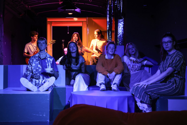 Fünf Jugendliche sitzen nebeneinander auf einer Art Couch. Hinter ihnen spielt eine Band. Die Band ist beleuchtet. Fünf Jugendliche sitzen nebeneinander auf einer Art Couch. Hinter ihnen spielt eine Band. Die Band ist beleuchtet.
