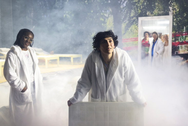 Zwei Schauspielerinnen stehen in weißen Bademänteln vorn an der Bühne. Hinter ihnen ist Nebel. In der hinteren Bühne kann man durch eine Tür weitere Schauspielerinnen sehen. Zwei Schauspielerinnen stehen in weißen Bademänteln vorn an der Bühne. Hinter ihnen ist Nebel. In der hinteren Bühne kann man durch eine Tür weitere Schauspielerinnen sehen.