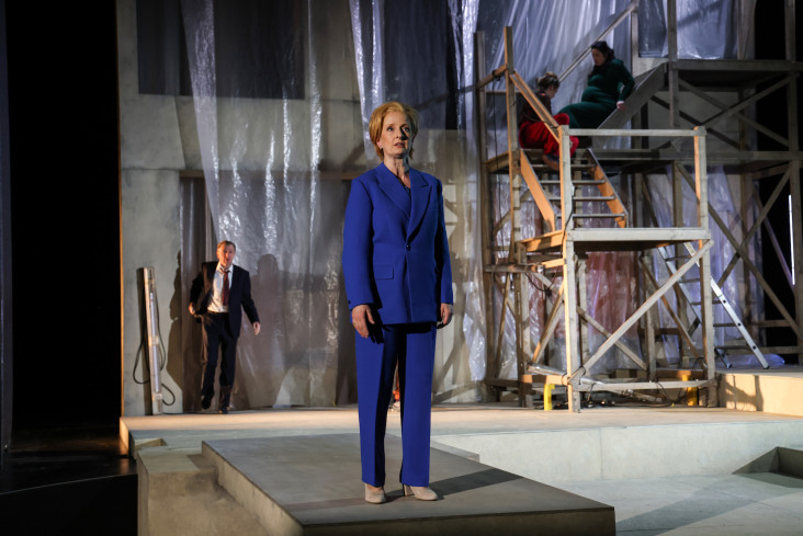Eine Schauspielerin steht vorn auf einer Rampe. Sie trägt einen blauen Hosenanzug. Hinten auf der Bühne kommt ein Schauspieler gelaufen. Er trägt einen dunklen Anzug.