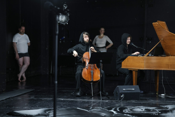 Ein Musiker spielt Cello, ein anderer sitzt am Flügel. Zwei Personen lehnen an der Wand.