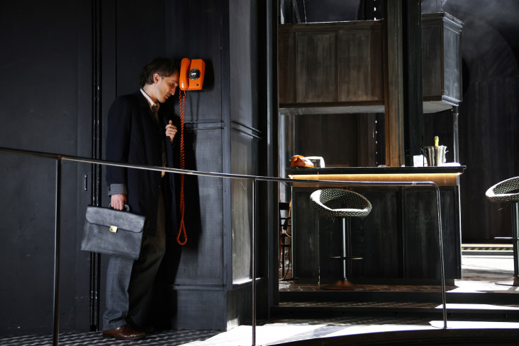 Neben einer Bar hängt ein orangenes Telefon an der Wand. Neben dem steht ein Mann in einem dunklen Mantel mit einer Aktentasche. Er lehnt die Stirn an das Telefon.