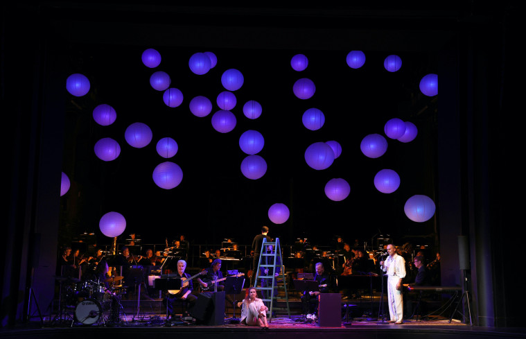 In der Mitte der Bühne sitzt eine Sängerin auf dem Boden, rechts neben ihr steht ein Mann und singt. Im Hintergrund spielt eine Band und das große Orchester. Vom Bühnenhimmel hängen sehr viele violett leuchtende Lampions. In der Mitte der Bühne sitzt eine Sängerin auf dem Boden, rechts neben ihr steht ein Mann und singt. Im Hintergrund spielt eine Band und das große Orchester. Vom Bühnenhimmel hängen sehr viele violett leuchtende Lampions.