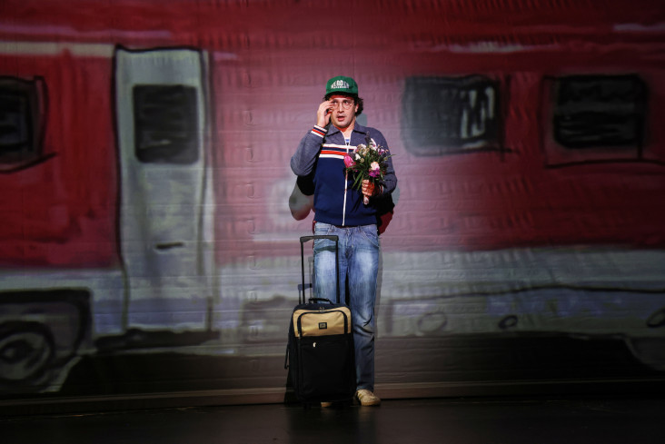Ein Schauspieler mit einem Rollkoffer steht auf der Bühne. Hinter ihm ist ein gemalter Zug. Ein Schauspieler mit einem Rollkoffer steht auf der Bühne. Hinter ihm ist ein gemalter Zug.