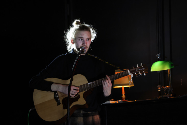 Ein Musiker mit Gitarre und Mikrofon. Neben ihm brennen zwei Schreibtischlampen.