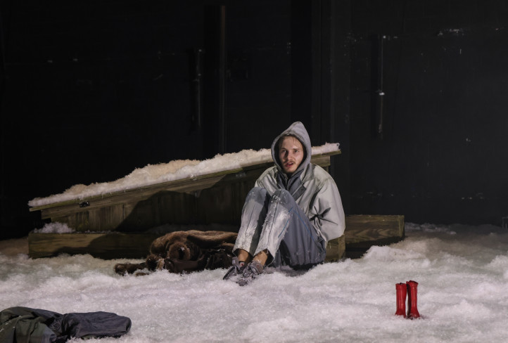 Ein Schauspieler sitzt mit angezogenen Beinen auf dem Boden. Er trägt eine Kapuze über dem Kopf. Um ihn herum liegt Kunstschnee. Neben ihm stehen zwei rote Kinderstiefel.