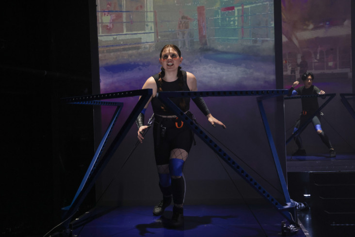 Eine Person steht vor einer Leinwand. Sie ist von blauen Stahlrahmen umgeben. Auf der Leinwand ist ein Boxring abgebildet.