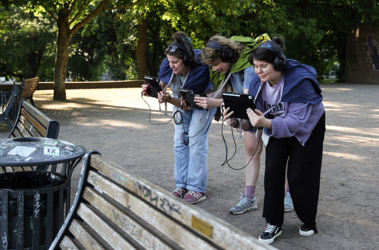 Drei Personen stehen leicht vorgebeugt nebeneinander. Alle tragen Kopfhörer. In den Händen halten sie Tablets. Ihnen gegenüber stehen zwei Bänke und ein Mülleimer. Sie sind in einem Park.