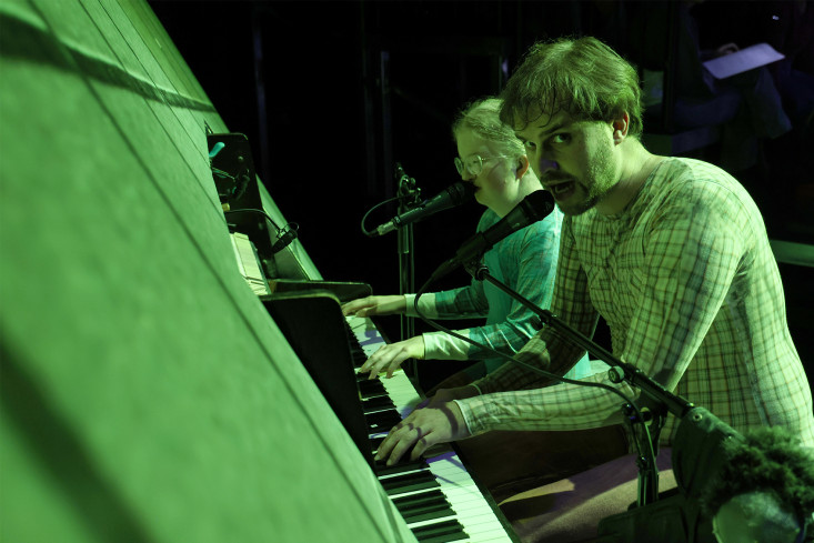 An einem Klavier sitzen zwei Personen. Sie singen in Mikrofone. An einem Klavier sitzen zwei Personen. Sie singen in Mikrofone.
