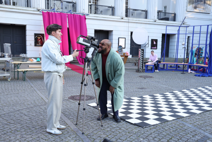 Ein Schauspieler filmt als Kameramann einen anderen Schauspieler. Der schaut ganz nah in die Kameralinse. An der Front des Theaters hängt ein großer, pinker Vorhang. Ein Schauspieler filmt als Kameramann einen anderen Schauspieler. Der schaut ganz nah in die Kameralinse. An der Front des Theaters hängt ein großer, pinker Vorhang.